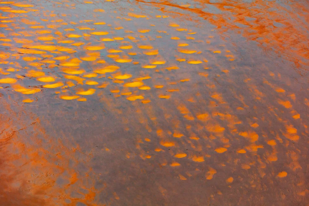 Shallow water with vibrant orange and yellow mineral deposits reflecting light, creating a rippled pattern across the wet sand.