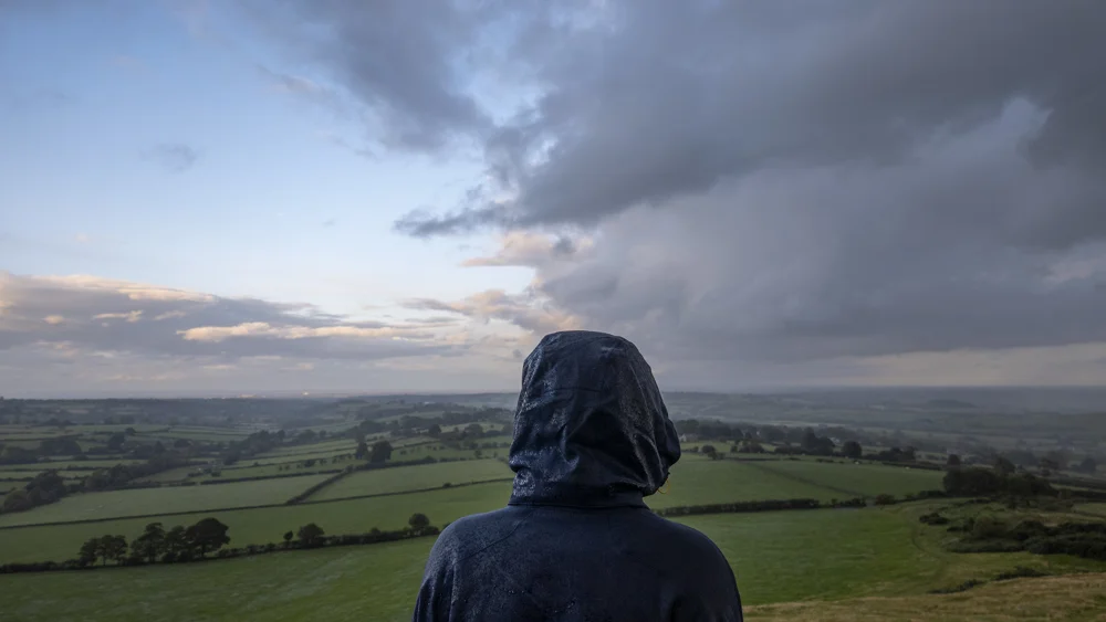 Hooded figure in a wet raincoat looking out over patchwork green fields from a hilltop at dusk under heavy cloud