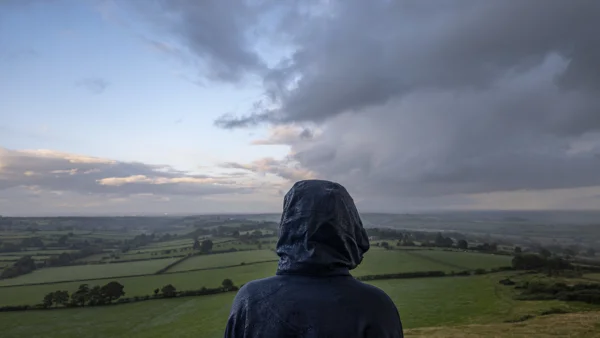 Hooded figure in a wet raincoat looking out over patchwork green fields from a hilltop at dusk under heavy cloud
