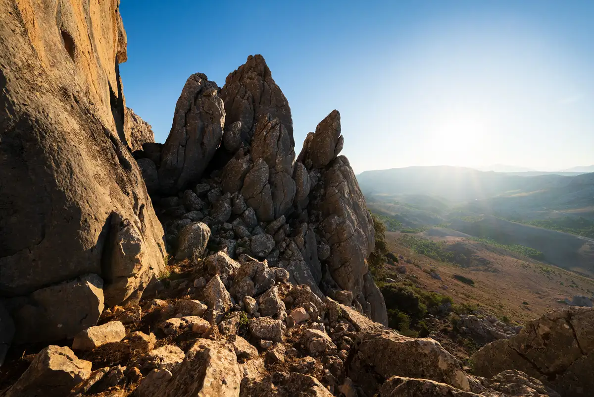 Rugged rocky outcrops sharply rise in the foreground, casting shadows in the warm light of the setting sun. The landscape extends into a vast valley with rolling hills and scattered greenery under a clear blue sky. Sunlight streams across the scene, creating a serene and expansive view.