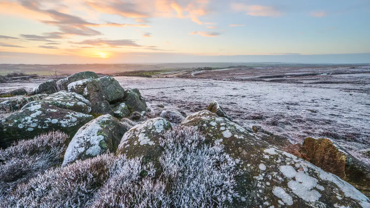 Frost-covered moorland at sunrise, with large, lichen-spotted boulders in the foreground. The landscape stretches towards the horizon, displaying patchy fields and distant hills under a sky painted with soft orange and blue hues.
