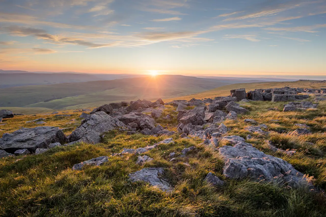 A panoramic view of a rocky landscape with scattered grey boulders and patches of grass, illuminated by the golden light of a setting sun on the horizon. The sky is painted with soft clouds, adding a serene atmosphere to the scene. Rolling hills extend into the distance under the vivid sky.