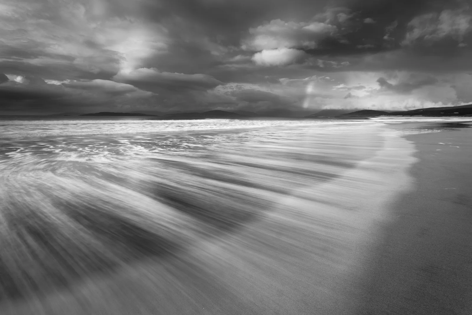 Monochrome seascape with long-exposure waves rolling onto a flat beach, distant hills visible across calm waters beneath dramatic cloudy sky.