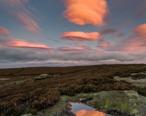 Moorland Landscapes: Lenticular clouds with a pinkish hue reflect the fading light. Below, the rugged heathland is subdued in shadow, but the sky's warm tones vividly mirror in a small puddle on the rocky ground, hinting at the sky's drama. a view of a rocky mountain