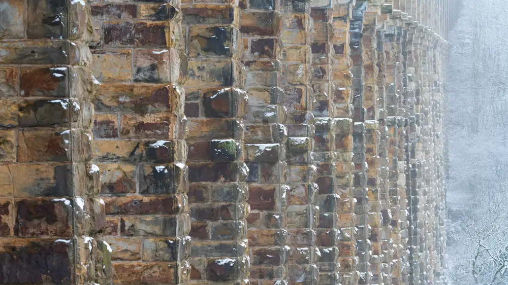 Close-up view of a snow-dusted stone bridge or viaduct showing textured, weathered stone blocks in a repeating pattern. The structure stretches into the distance on the right, with a snowy forest visible in the background, partially obscured by mist.