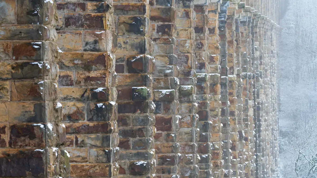 Close-up view of a snow-dusted stone bridge or viaduct showing textured, weathered stone blocks in a repeating pattern. The structure stretches into the distance on the right, with a snowy forest visible in the background, partially obscured by mist.