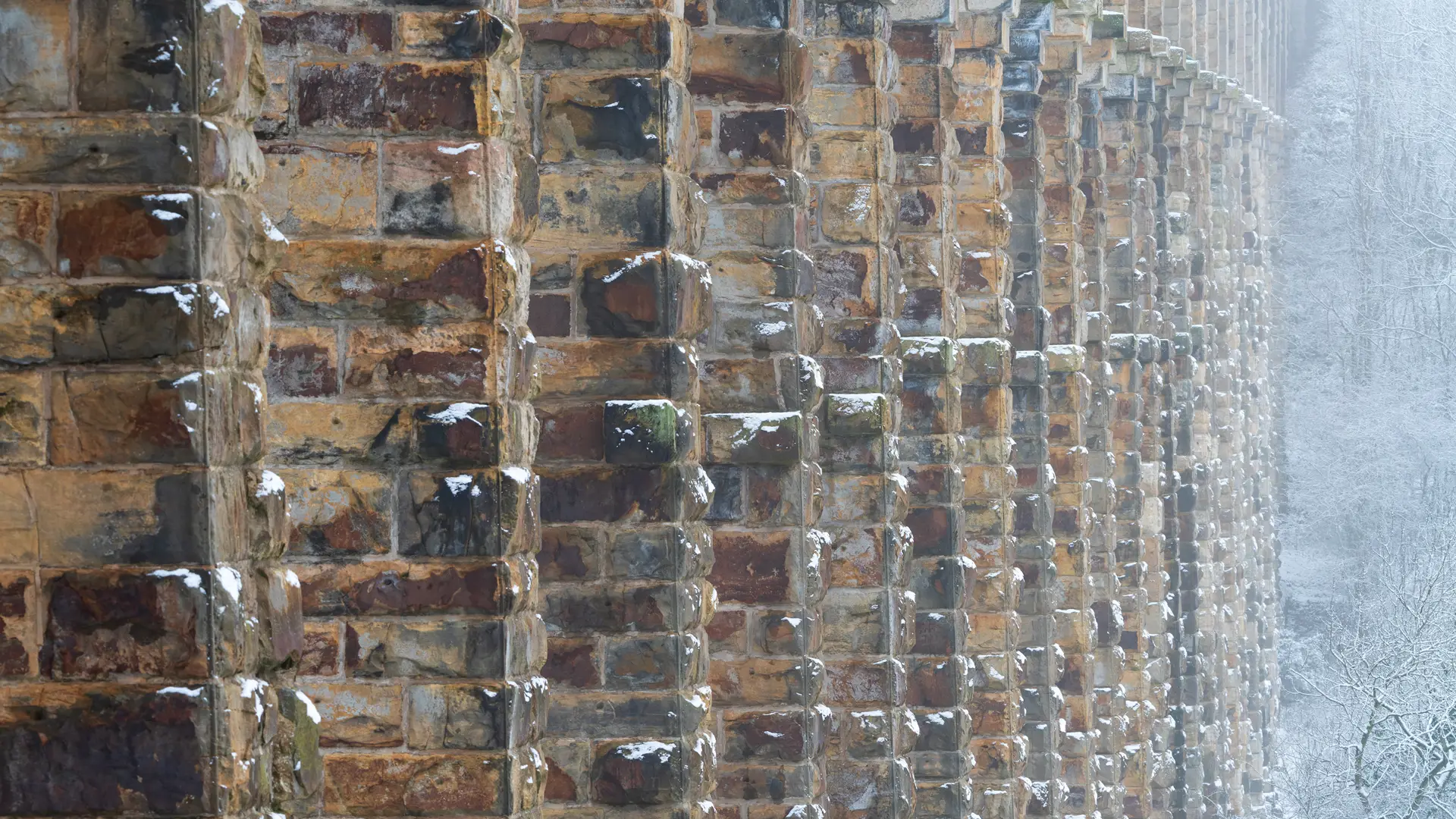 Close-up view of a snow-dusted stone bridge or viaduct showing textured, weathered stone blocks in a repeating pattern. The structure stretches into the distance on the right, with a snowy forest visible in the background, partially obscured by mist.