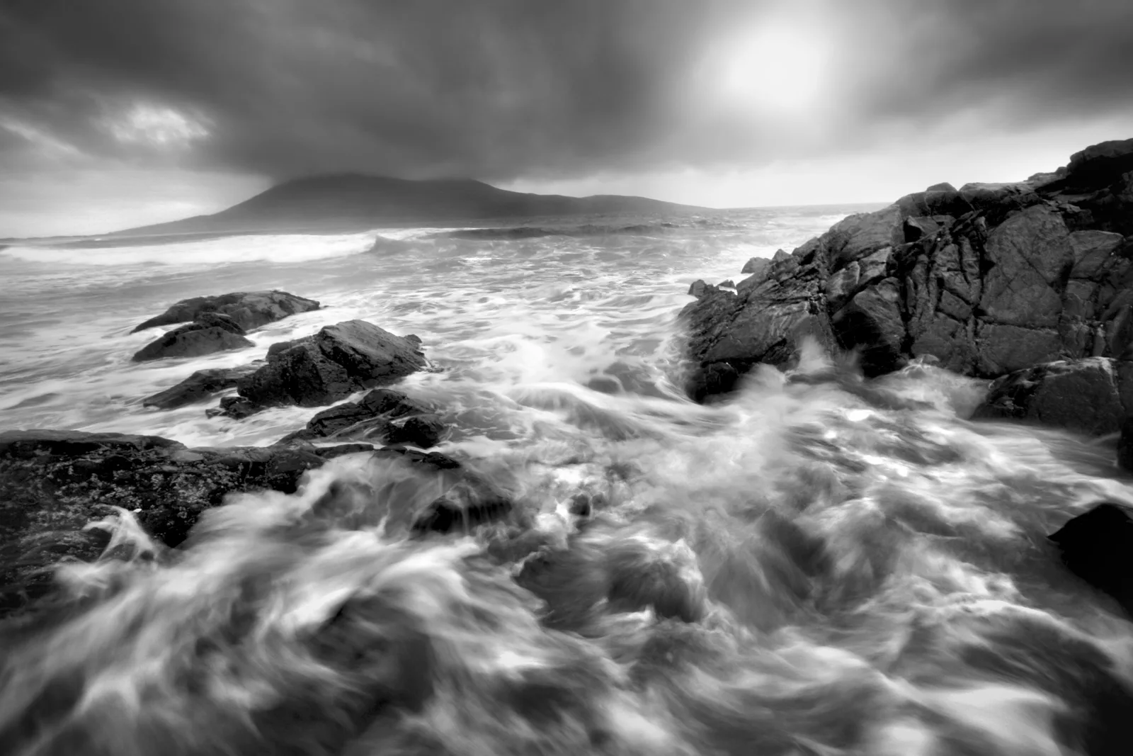 Monochrome coastal scene with rocky foreground, turbulent waves, and a conical mountain across the bay under stormy skies.