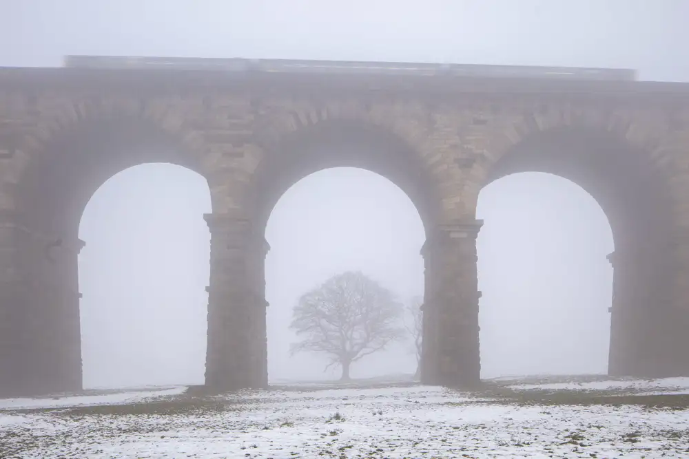A stone viaduct with three large arches stands in a foggy landscape. A train is faintly visible on top, blurred by mist. Below, a solitary leafless tree is seen through the central arch. The ground is covered in patches of snow, adding to the wintry atmosphere.