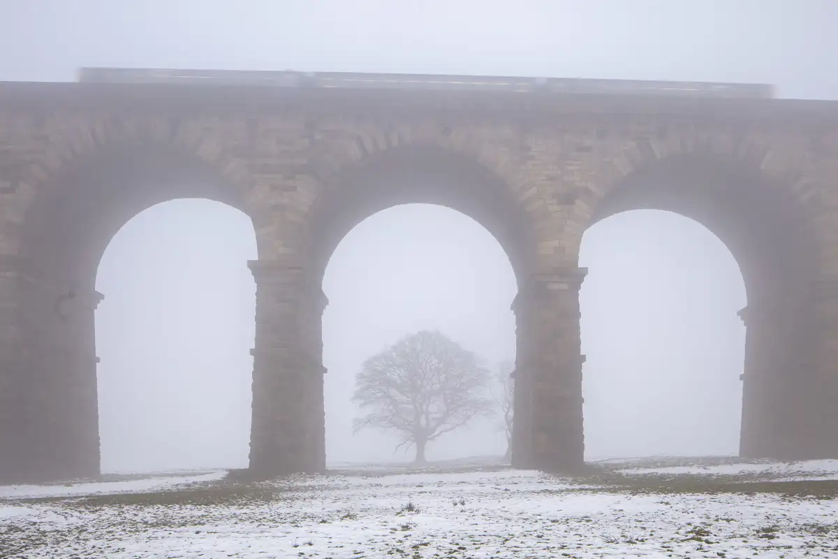 A stone viaduct with three large arches stands in a foggy landscape. A train is faintly visible on top, blurred by mist. Below, a solitary leafless tree is seen through the central arch. The ground is covered in patches of snow, adding to the wintry atmosphere.