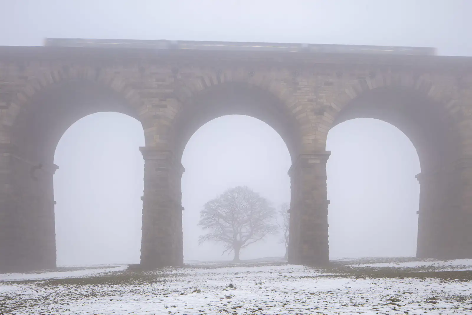 A stone viaduct with three large arches stands in a foggy landscape. A train is faintly visible on top, blurred by mist. Below, a solitary leafless tree is seen through the central arch. The ground is covered in patches of snow, adding to the wintry atmosphere.