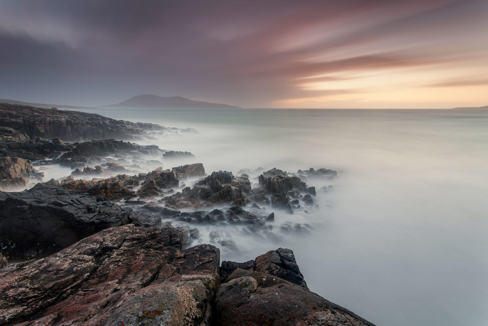 Rocky coastal landscape with jagged black rocks extending into the misty, blurred sea. A mountain is visible in the hazy background beneath a dramatic sky with shades of purple and orange, indicating sunrise or sunset.