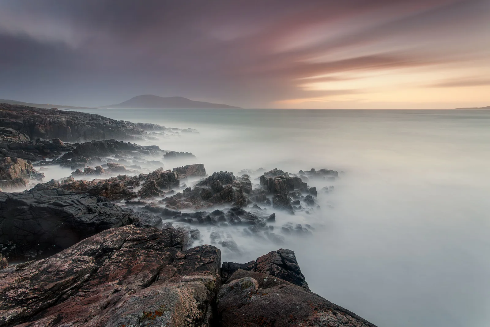 Rocky coastline at sunset with misty sea, distant mountain across calm waters under soft pink and purple sky.