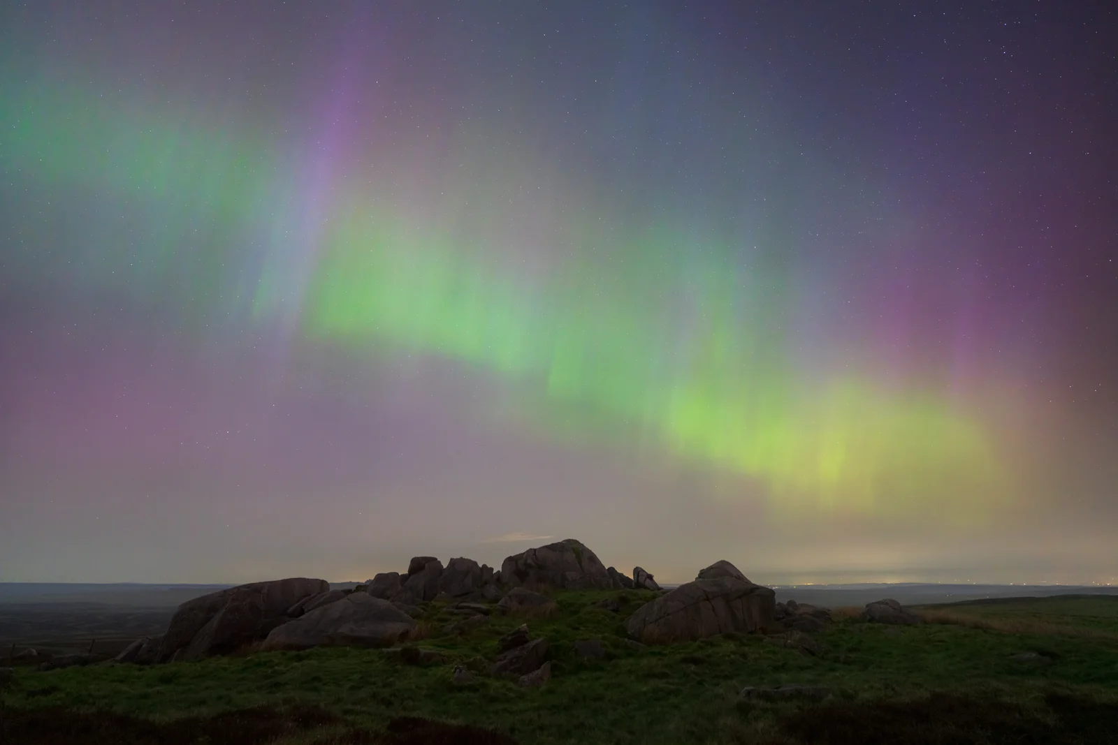 Northern lights displaying vibrant green and purple hues across the night sky, above a rocky landscape with grassy patches in the foreground. Stars are visible in the clear sky.