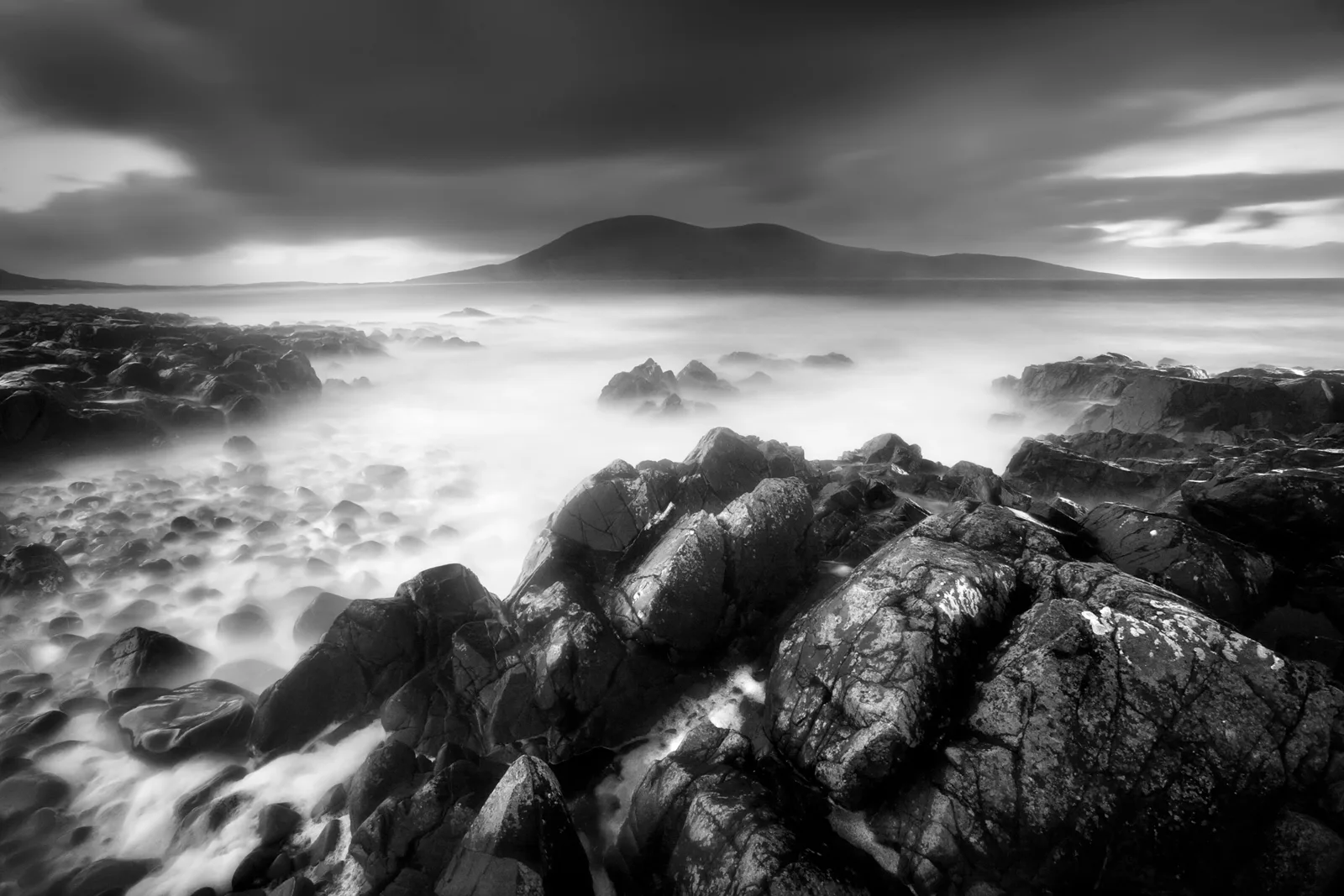 Rocky coastline with misty sea and distant mountain in monochrome, long-exposure photograph.