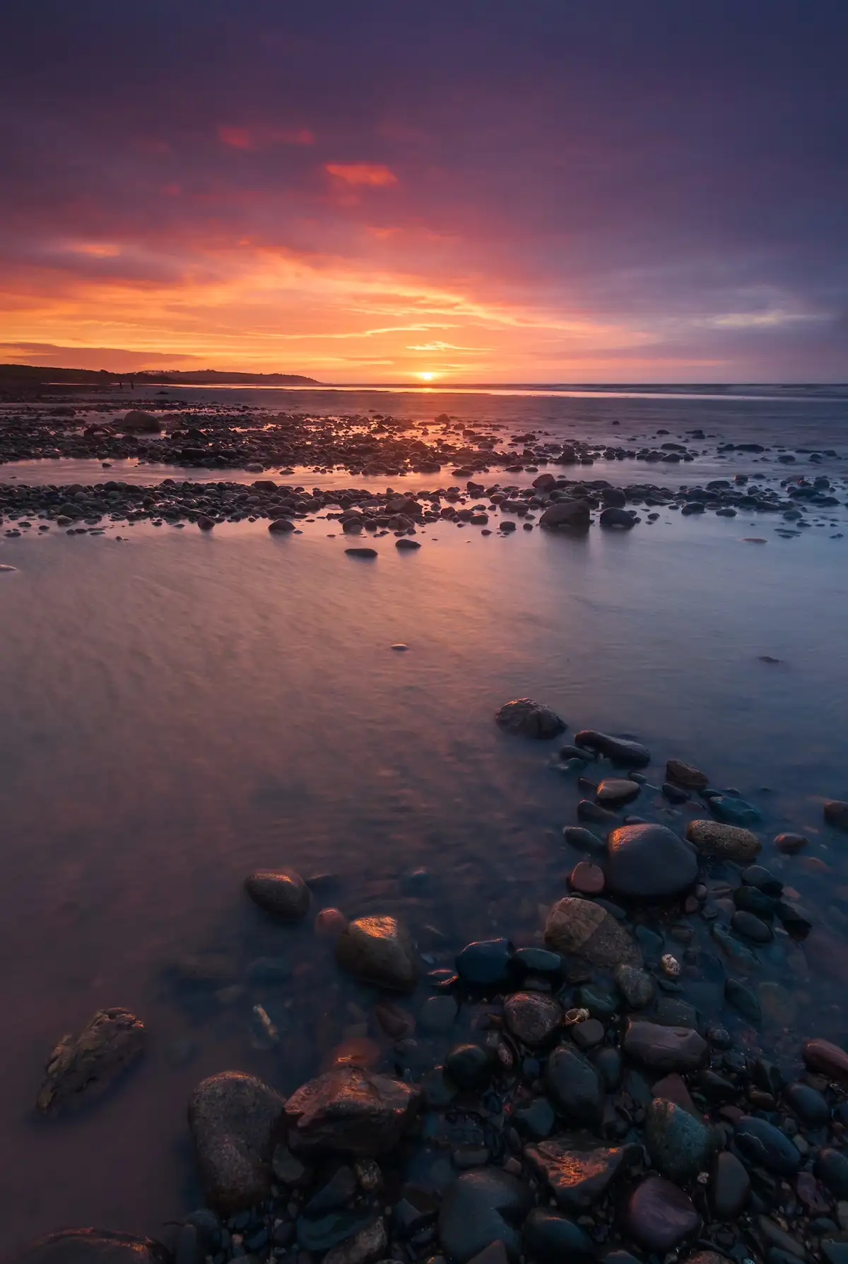 Sunset over a rocky shoreline with smooth stones scattered across the foreground. The sky is vibrant, filled with shades of orange, pink, and purple, reflecting on the water. The horizon features a gentle silhouette of distant land.