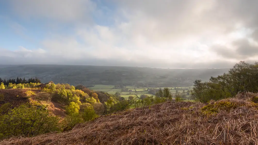 Overlooking a wide valley, with a blend of sunlit and shadowed areas. The foreground features dry, brown bracken and green trees, leading into lush, green fields below. The horizon is marked by hazy hills under a sky with scattered clouds and blue patches.