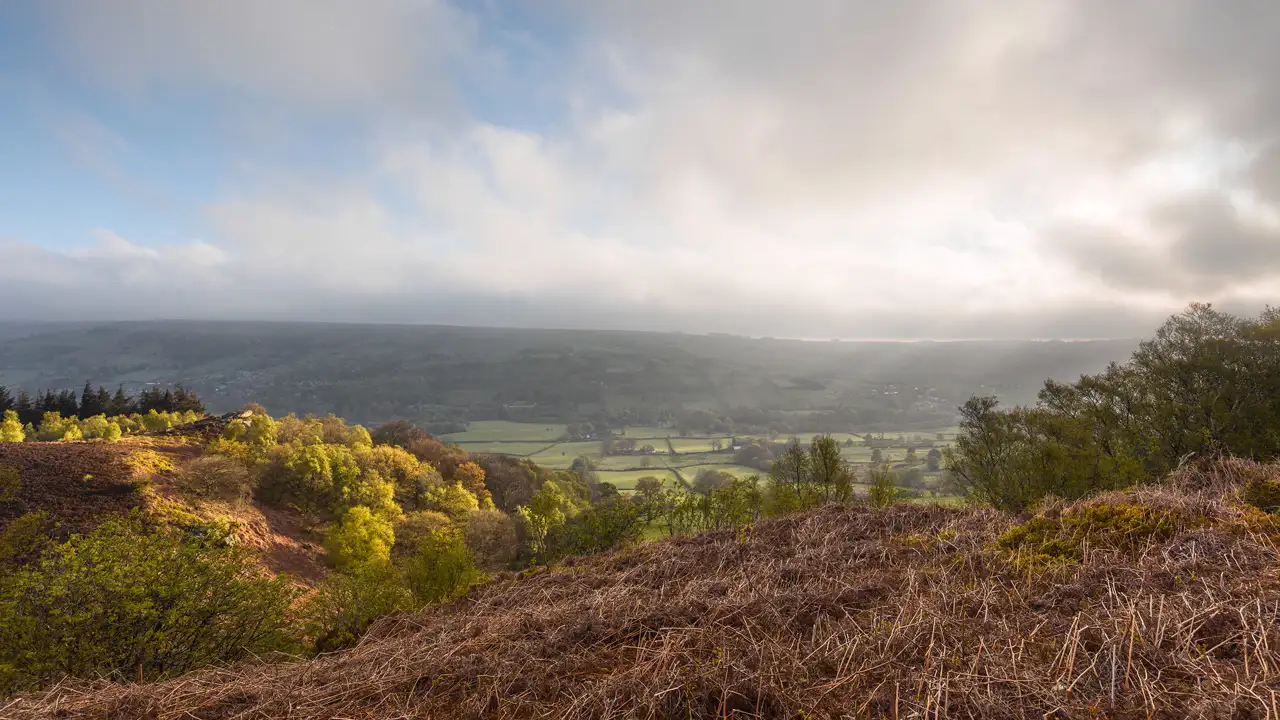 Overlooking a wide valley, with a blend of sunlit and shadowed areas. The foreground features dry, brown bracken and green trees, leading into lush, green fields below. The horizon is marked by hazy hills under a sky with scattered clouds and blue patches.